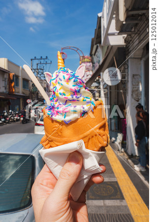 A whimsical unicorn themed ice cream in a fish shaped waffle cone held against a sunny urban street in Phuket, Thailand, with motorbikes and shop signs. 125012014