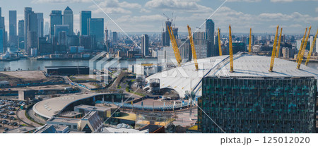Aerial perspective of London with the O2 Arena's white dome and yellow supports in the foreground, Canary Wharf skyscrapers, and the River Thames. 125012020