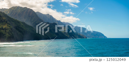 Towering green cliffs of the Napali Coast meet the vibrant blue ocean under a bright sky with scattered clouds, illuminated by soft sunlight. 125012049
