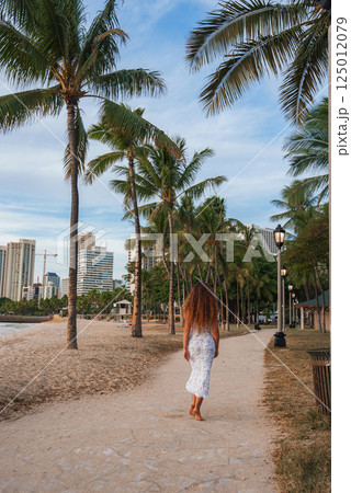 A woman in a white dress walks along a sandy path near a beach in Honolulu, Hawaii, with palm trees, ocean views, and high rise buildings in the background. 125012079