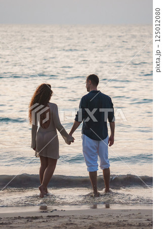 A couple stands barefoot at the water's edge on a tranquil beach, facing the calm sea during soft lighting, likely in Phuket or Koh Phi Phi, Thailand. A couple stands barefoot at the water's edge on a tranquil beach, facing the calm sea during soft lighting, likely in Phuket or Koh Phi Phi, Thailand. 125012080