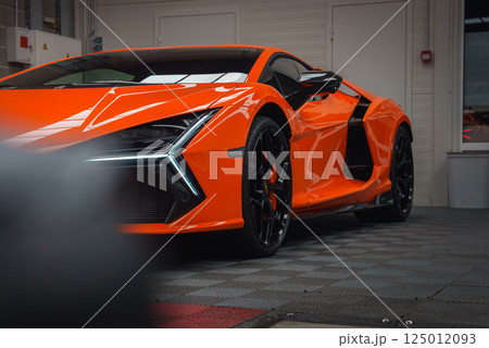 An orange Lamborghini Revuelto is displayed indoors, highlighting its sleek design, modern LED headlights, and glossy finish on tiled flooring. 125012093