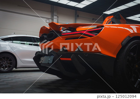 The image features the rear end of an orange Lamborghini Revuelto, highlighting its taillights and dual exhausts. A white car is visible in the background. 125012094