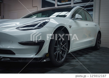 A white Tesla Model 3 is parked indoors on a checkered floor, highlighting its sleek design, aerodynamic lines, and distinctive front headlights. 125012187