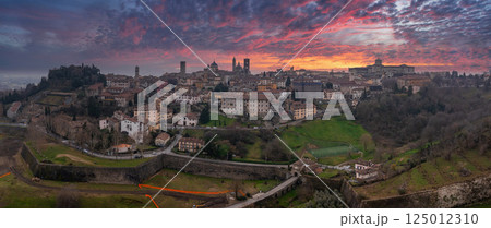 The old town of Bergamo, Italy, features medieval towers, domes, and Venetian walls under a vibrant sunset with pink, orange, and purple hues. The old town of Bergamo, Italy, features medieval towers, domes, and Venetian walls under a vibrant sunset with pink, orange, and purple hues. 125012310