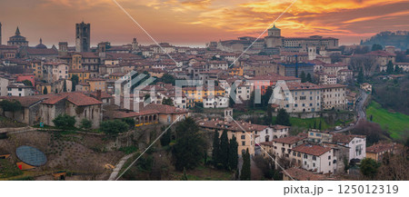 The old town of Bergamo, Italy, features Torre del Gombito, the Basilica of Santa Maria Maggiore, terracotta rooftops, and rolling hills at sunset. 125012319
