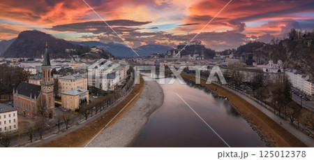 Aerial view of Salzburg, Austria, at sunset featuring the Salzach River, historic buildings, Hohensalzburg Fortress, and a vibrant orange and pink sky. 125012378