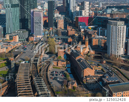Aerial view of Manchester, UK, showing modern skyscrapers like Beetham Tower, historic red brick buildings, railway tracks, and canals on a sunny day. 125012428