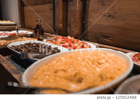 A buffet featuring scrambled eggs, mushrooms, tomatoes, and cured meats in a luxury Alpine hotel. Wooden backdrop and warm lighting create a cozy ambiance. 125012488