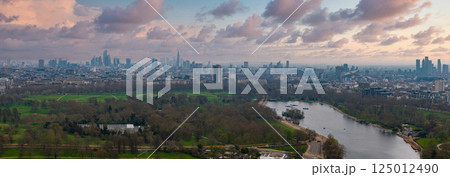 Aerial perspective of London featuring The Shard, Canary Wharf, a green park with a winding river, and the city's urban sprawl under an overcast sky. 125012490