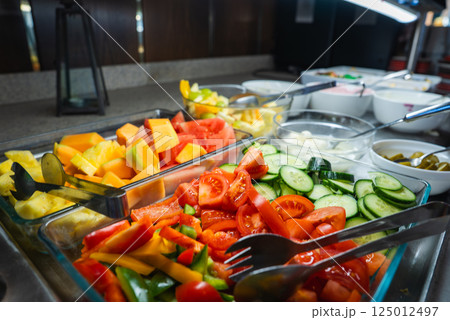 An indoor buffet setup features watermelon, pineapple, cantaloupe, tomatoes, cucumbers, and bell peppers in glass trays with serving tongs on a polished countertop. 125012497