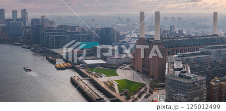 Aerial view of Battersea Power Station in London with its four chimneys, modern buildings, a green park, and the River Thames with a boat visible. 125012499