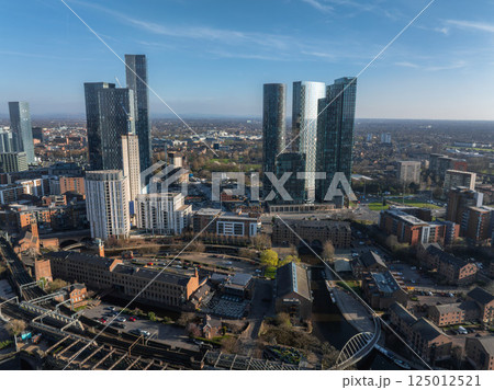 Aerial view of Manchester, UK, showing modern skyscrapers like Deansgate Square, historic brick buildings, canals, and suburban areas under a clear sky. 125012521