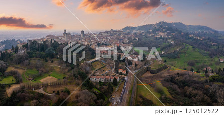 The old town of Bergamo, Italy, features medieval towers, domes, and Venetian walls, framed by green hills and bathed in warm sunset hues. 125012522