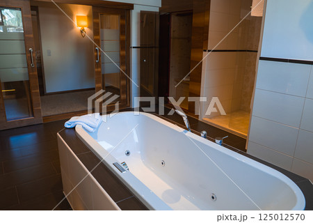 Bathroom interior with a white bathtub, dark flooring, tiled walls, wooden sliding doors, and soft lighting in a luxury hotel in Ischgl, Austria. Bathroom interior with a white bathtub, dark flooring, tiled walls, wooden sliding doors, and soft lighting in a luxury hotel in Ischgl, Austria. 125012570