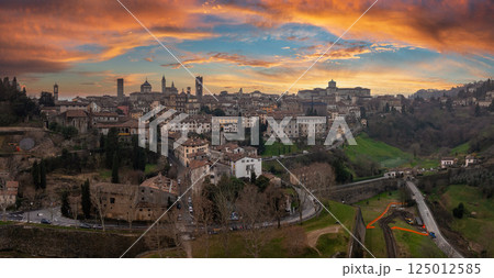 The old town of Bergamo, Italy, features medieval towers, domes, stone walls, and winding roads, set against a vibrant orange and pink sunset. The old town of Bergamo, Italy, features medieval towers, domes, stone walls, and winding roads, set against a vibrant orange and pink sunset. 125012585