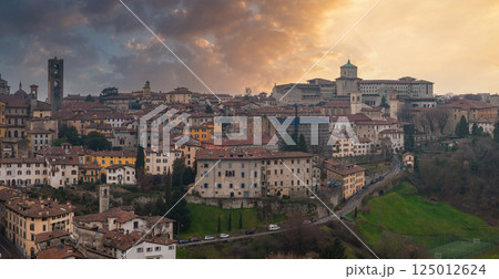 View of Bergamo's old town in Italy during sunset, featuring terracotta roofs, a bell tower, a green domed church, and a lush green hillside. 125012624