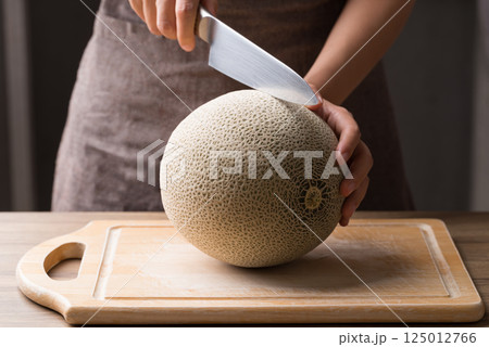 Hand holding kitchen knife and cutting melon fruit on wooden board 125012766