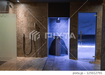 Spa area featuring stone walls, modern showers, a blue lit stone basin, and a glass door leading to a steam sauna in a high end Italian Alps hotel. 125012791