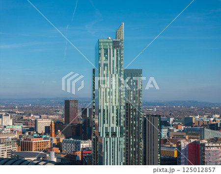 Aerial view of Manchester, UK, showcasing a sleek glass skyscraper with angular design, surrounded by diverse architecture and a sprawling urban landscape. 125012857