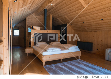 Wooden interior of an alpine hotel room with sloped ceiling, modern double bed, bedside table, radiator, towel warmer, and natural light from a window. Wooden interior of an alpine hotel room with sloped ceiling, modern double bed, bedside table, radiator, towel warmer, and natural light from a window. 125012974