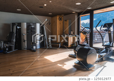 A gym with large windows showcasing the Italian Alps, featuring high end fitness equipment, wooden accents, and natural light enhancing the alpine setting. 125012977