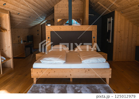 A cozy hotel room with natural wood paneling, a large wooden bed, built in nightstands, a sloped ceiling, and natural light in the Italian Alps. A cozy hotel room with natural wood paneling, a large wooden bed, built in nightstands, a sloped ceiling, and natural light in the Italian Alps. 125012979