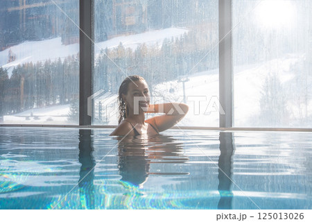 A woman enjoys an indoor infinity pool with floor to ceiling windows overlooking snowy slopes, evergreen trees, and soft sunlight in the Italian Alps. 125013026