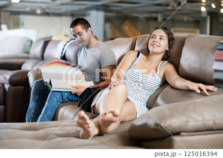 In store wife settled down with legs to test of sofa in use,husband examines upholstery fabric In store wife settled down with legs to test of sofa in use,husband examines upholstery fabric 125016394
