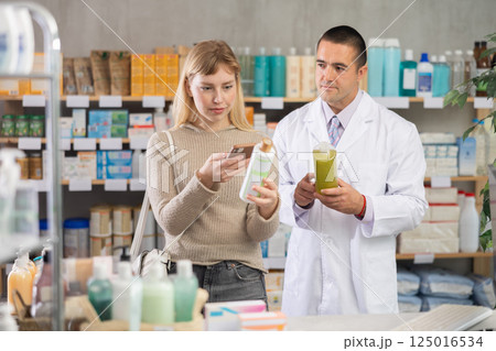 Young woman scanning QR-code on shampoo while pharmacist offering Young woman scanning QR-code on shampoo while pharmacist offering 125016534