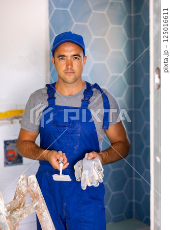 Professional man builder in blue construction overalls and cap posing with gloves and trowel in apartment Professional man builder in blue construction overalls and cap posing with gloves and trowel in apartment 125016611