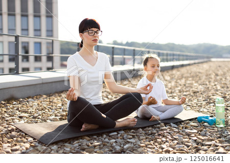 Mother and daughter practicing yoga on rooftop. Young woman and girl meditating outdoors, serene atmosphere. Family bond, yoga lifestyle, healthy living. 125016641
