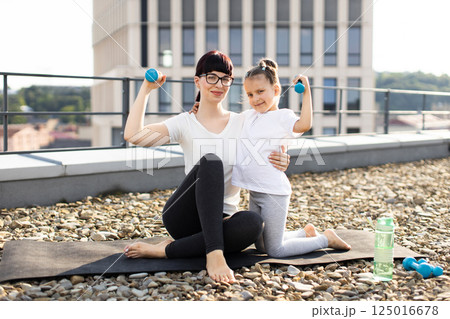 Young adult woman and daughter practicing fitness with dumbbells outdoors on rooftop terrace, showcasing healthy lifestyle concept. Young adult woman and daughter practicing fitness with dumbbells outdoors on rooftop terrace, showcasing healthy lifestyle concept. 125016678