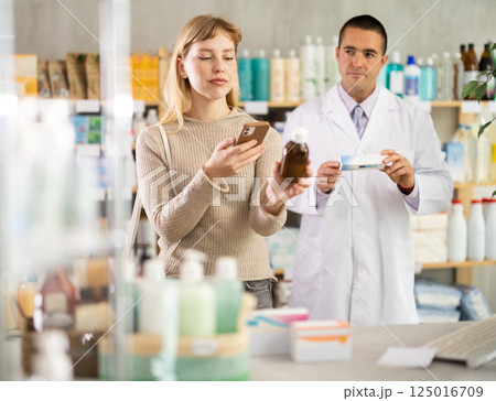 Young woman in pharmacy scans QR code on antitussive syrup, male pharmacist in background 125016709