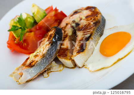 Grilled salmon with fried egg, tomatoes, avocado on white dish 125016795