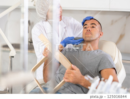 Female doctor, together with male patient, examines face in mirror after undergoing hardware procedure for skin rejuvenation 125016844
