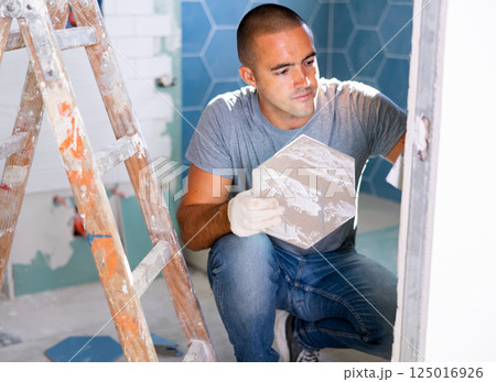 The technology of laying tile, repair and decoration. Focused man puts porcelain tiles on wall sitting squat at bathroom 125016926