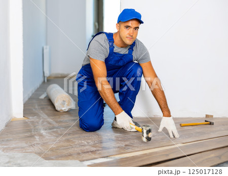 Professional man builder in blue construction overalls laying laminate flooring by rubber hammer Professional man builder in blue construction overalls laying laminate flooring by rubber hammer 125017128