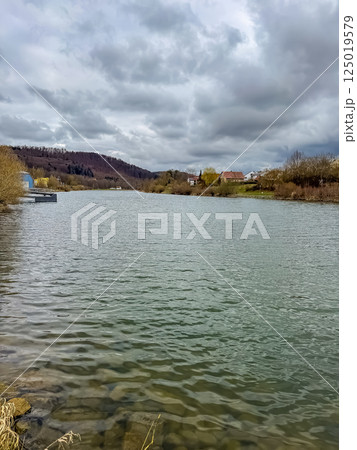 Tranquil river with cloudy sky and hills in distance, rural village shoreline. High quality photo Tranquil river with cloudy sky and hills in distance, rural village shoreline. High quality photo 125019579