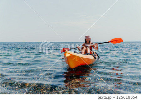 Woman Kayak Paddle Ocean - A woman in a swimsuit and a hat paddles an orange kayak in the sea on a bright, clear day. 125019654