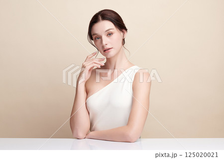 A beautiful young woman with clean, fresh skin against a beige background. Skincare, facial treatment, cosmetology, beauty, and spa. An elegant portrait of an woman. 125020021
