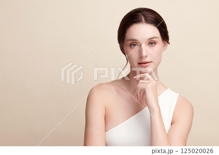A beautiful young woman with clean, fresh skin against a beige background. Skincare, facial treatment, cosmetology, beauty, and spa. An elegant portrait of an woman. 125020026