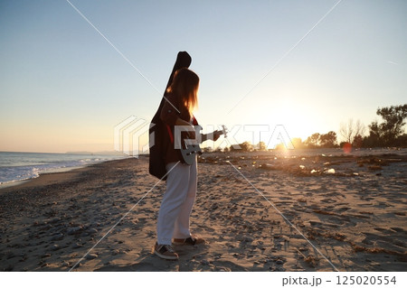 Girl Walks And Plays The Bass Instrument On The Beach Near The Ocean At Sunset Girl Walks And Plays The Bass Instrument On The Beach Near The Ocean At Sunset 125020554
