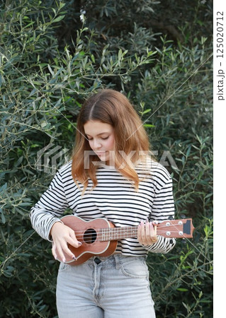 Girl Strums With Ukulele Musical Instrument Near A Tree In The Mountains 125020712