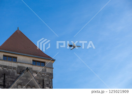 Airplane flying over historic stone building with red tiled roof against bright blue sky Airplane flying over historic stone building with red tiled roof against bright blue sky 125021909