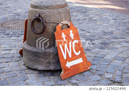 Handmade wooden WC sign with male and female icons and arrow leaning against stone post on old cobblestone street Handmade wooden WC sign with male and female icons and arrow leaning against stone post on old cobblestone street 125022490