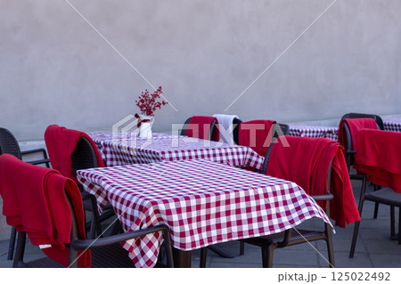 Outdoor cafe tables with red and white checkered tablecloths and blankets on chairs against plain wall on quiet city street 125022492