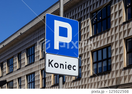 End of parking zone traffic sign on pole with blue background and modern building facade in sunlight under clear sky 125022508