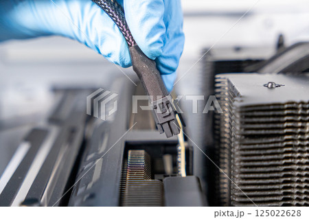 Technician in blue gloves holding 12VHPWR PCIe power cable above graphics card slot during PC assembly or GPU installation process 125022628