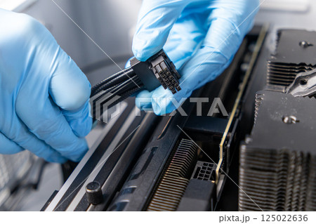 Technician wearing blue gloves connecting black power cable to graphics card inside high-performance desktop PC during hardware installation 125022636